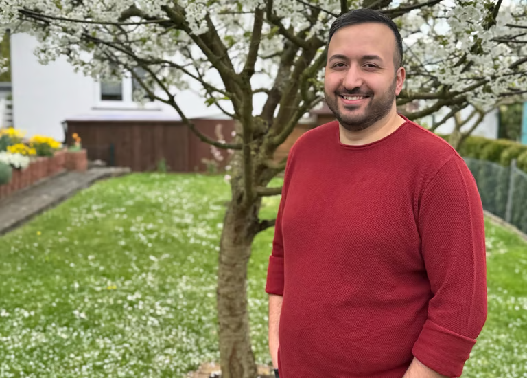 Mann mit kurzem Bart und rotem Pullover steht neben einem blühenden Baum im Garten.