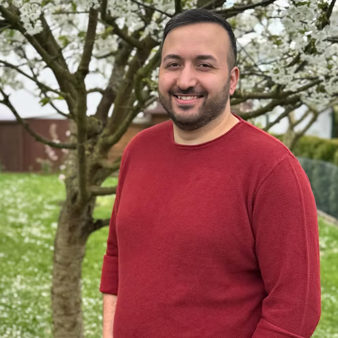Mann mit Bart trägt einen roten Pullover und steht vor einem blühenden Baum im Garten.